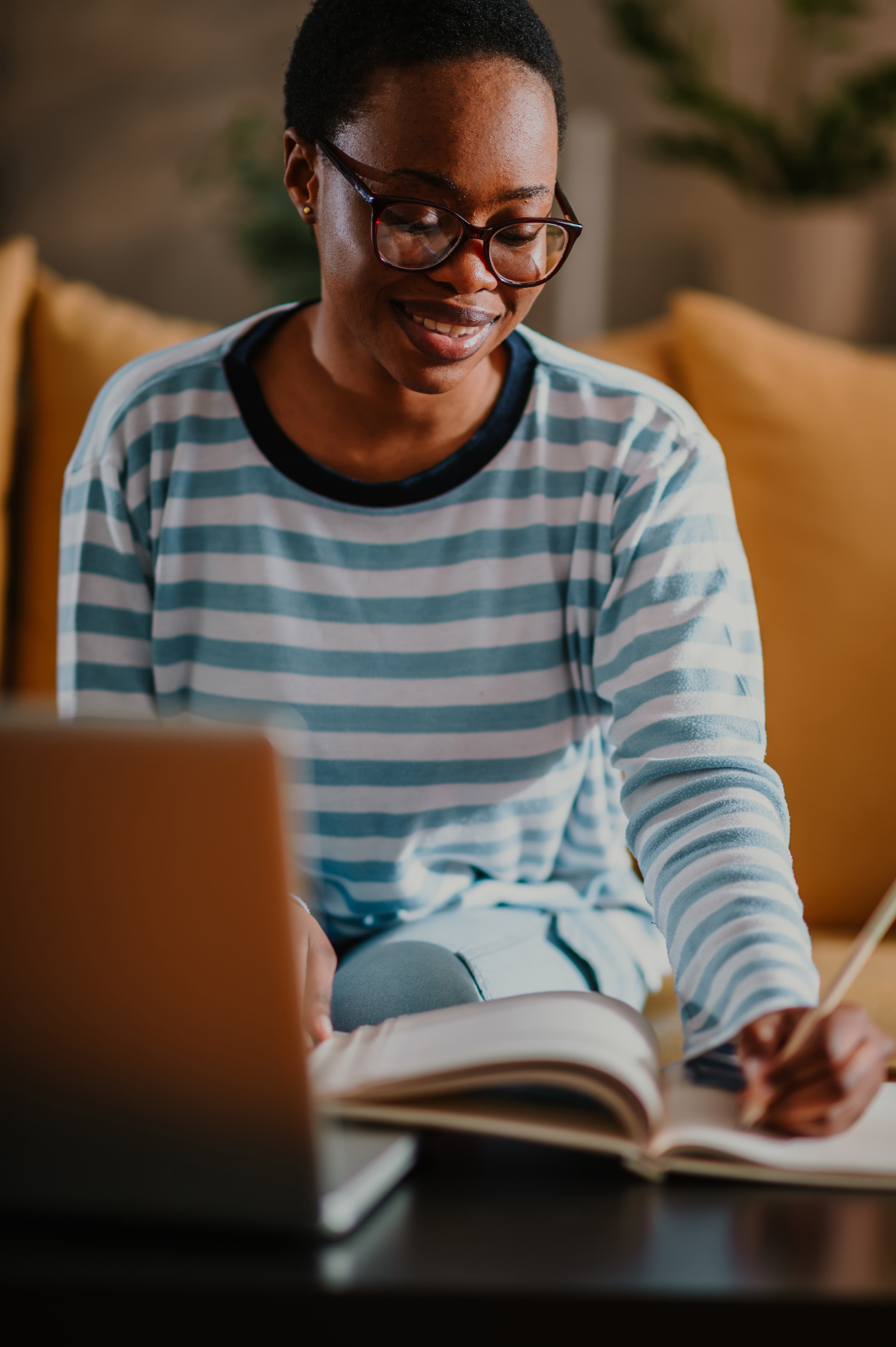 A smiling learner writing in a notebook at home with a laptop nearby